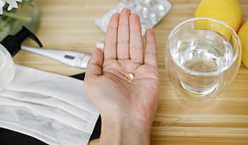 Vitamin D supplement in hand with water, lemon, thermometer, and mask on table