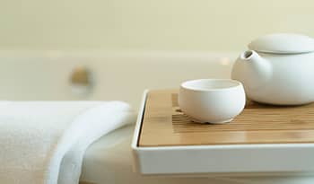 white teapot and teacup on a wooden tray next to a towel resting on the edge of a bathtub