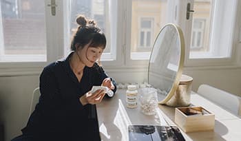 Woman doing skincare routine at vanity with supplements and beauty products on table