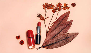 lipstick and lipgloss with some fall leaves sitting on a peach background
