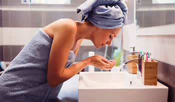 woman in bath towel washing face at sink with oil cleanser