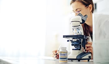 Researcher in lab looking through microscope with omega-3 supplement on table