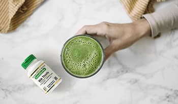 Cup of green juice in hand over marble table with milk thistle supplement bottle