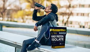 Man taking a break from a workout outside sitting on a bench drinking from sports bottle with bag of