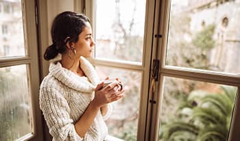 Young woman in sweater holding a cup of tea looks out the window