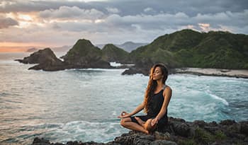 Woman practicing yoga sitting in lotus pose at sunset with on the coast with mountain view.