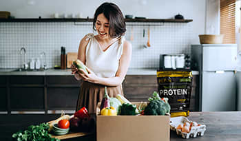 Asian woman unpacking healthy groceries in kitchen with vegan protein powder, vegetables, and eggs o
