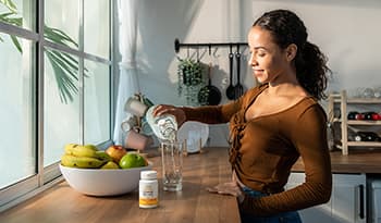 Young woman pouring glass of water in kitchen with vitamin K+D supplement on counter