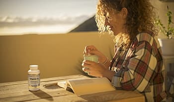 Woman sitting outside on her deck with coffee, a book, and vitamin D bottle